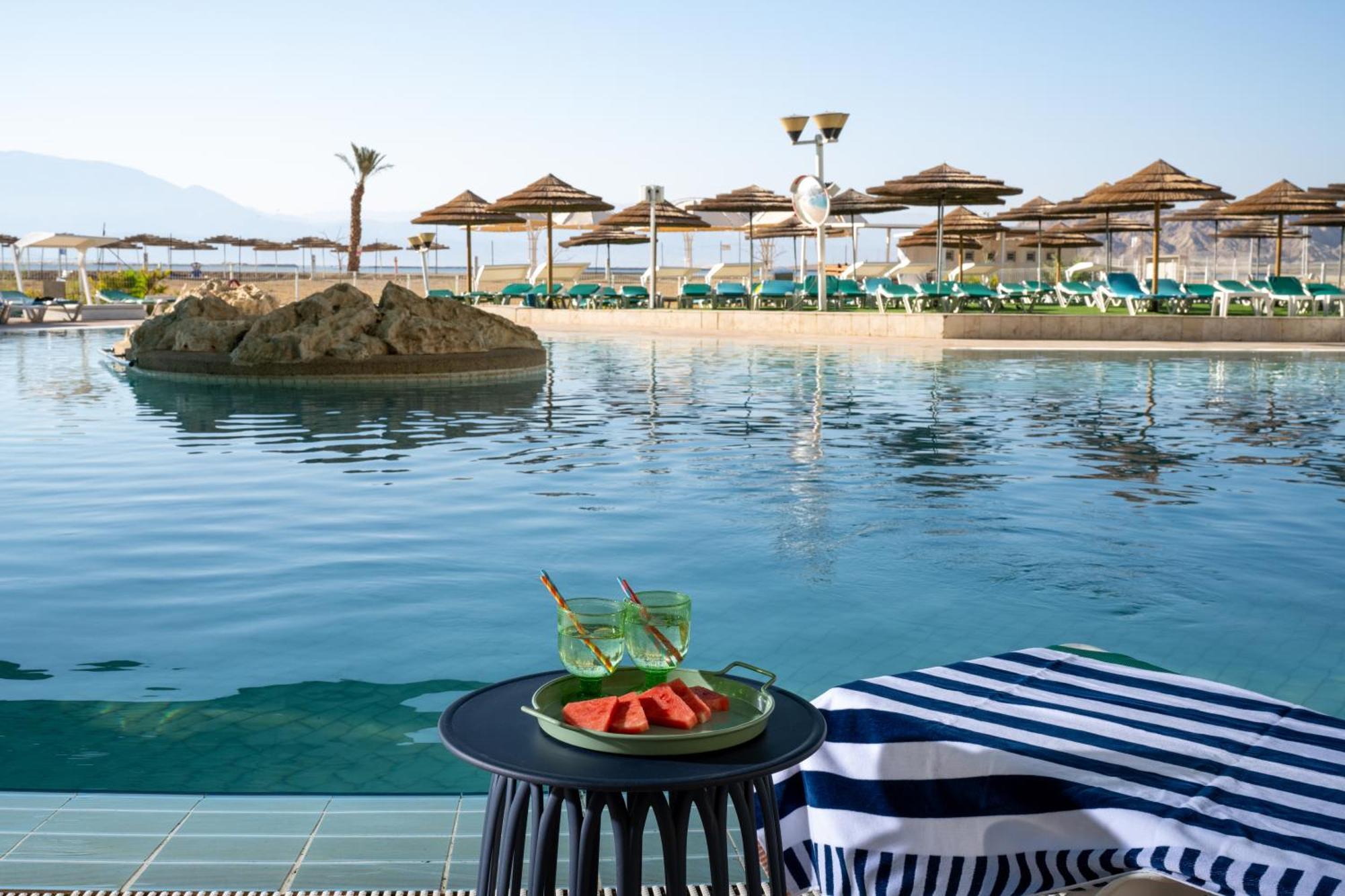 Leonardo Club Hotel Dead Sea Неве-Зогар Екстер'єр фото The photo depicts a tranquil swimming pool area at a resort or hotel. In the foreground, there is a small round table with a plate containing slices of watermelon and two green drinks. The background features a calm pool with a small rock island in t