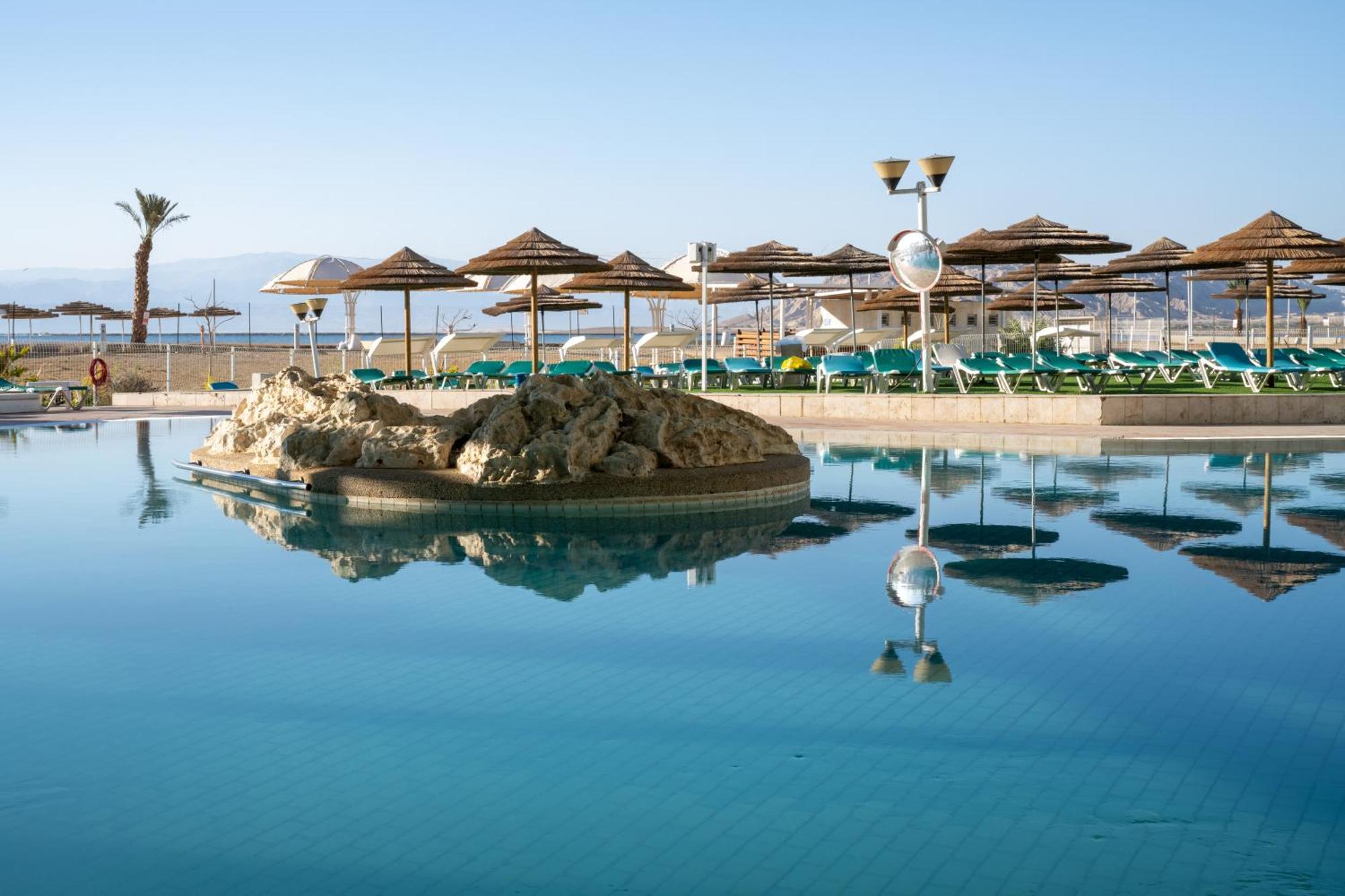 Leonardo Club Hotel Dead Sea Неве-Зогар Екстер'єр фото The photo shows a tranquil swimming pool area. It features a large, reflective pool with a small rock formation in the center. Surrounding the pool are several sun loungers with green cushions and a series of thatched umbrellas providing shade. In th